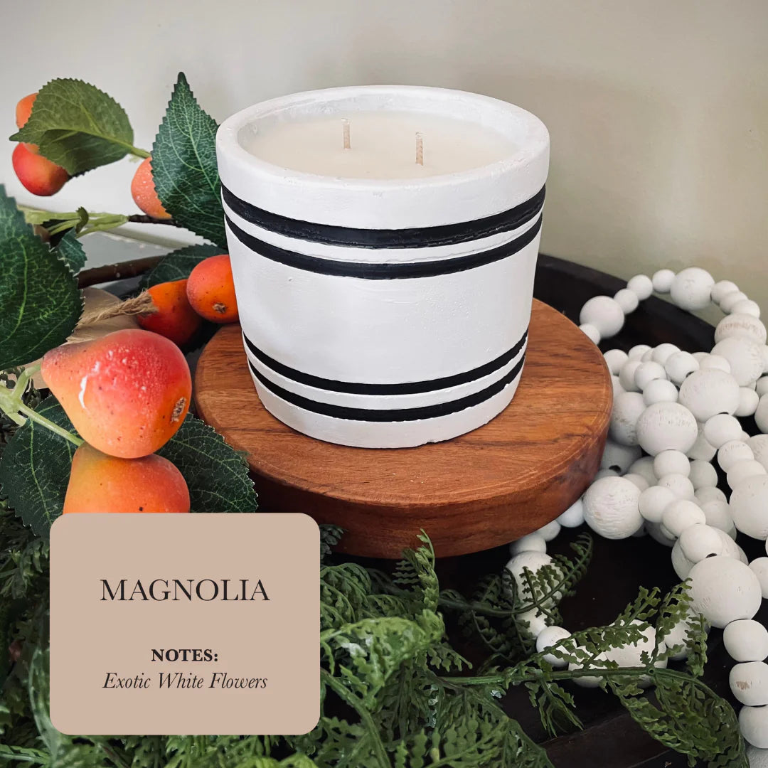A hand poured concrete candle titled 'Magnolia' by Blooming Violets, surrounded by fresh fruit and greenery, perfect for home decor with long lasting, fragrant qualities. Ideal for spring and summer settings, this elegant candle adds a touch of sophistication to any decor. Enjoy free shipping on this beautiful addition to your space.